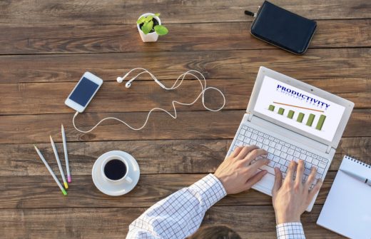 Natural Rough Wooden Desk and Man Working on Computer Top View Smart Casual Clothing Typing on Keyboard with Productivity Chart on Screen Smart Phone and Cup of Coffee aside